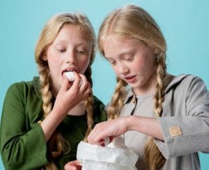 Two girls with blonde hair eat marshmallows out of a paper bag.
